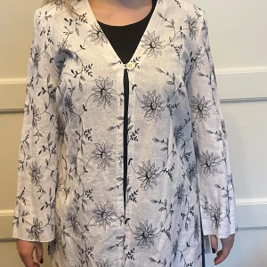Sommarfräsch Jacka/kavaj linne - Ofodrad lång blus/jacka i vitt linne med svart maskinbroderat blommönster, tunna axelvaddar, en knapp fram. Tvättas i 30 grader. Passar till svart topp av samma märke som annonseras av mig. Mycket fint skick, använd ett fåtal gånger.