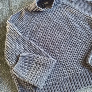 Grå stickad tröja - En varm och bekväm grå stickad tröja med ett sammetsliknande texturerat mönster och material. Tröjan har långa ärmar och ribbstickade muddar vid ärmslut, nederkant och halsringning. 