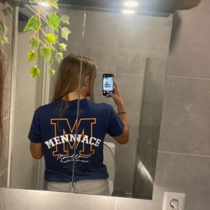 Blå t-shirt från Mennace - En blå t-shirt från Mennace med ett stort gult 'M' och texten 'MENNACE' i vitt på baksidan. T-shirten har korta ärmar och en normal passform.