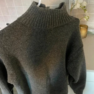 Grå stickad polotröja - En varm och bekväm grå stickad polotröja med ribbstickad krage. Tröjan har långa ärmar och en lös passform med detaljer såsom slits vid sidorna, vid kragen och i armarna, perfekt för kyligare väder.