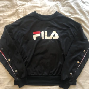 FILA tröja i nyskick st S - Svart med tryckknappar. Fina detaljer med rött och text Fila. Super snygg och knappt använd. Dottern skutta i längden så den är lite liten nu. Tröjan är i blankt material.  Denna långärmade Fila tröja mycket dyr i inköp.  Nypris 849:-  