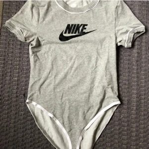 Grå NikeBody med svart text - Grå bodysuit från Nike, original, köpt för 399kr säljer för 170kr. Säljer den första bilden med grå body och svart Nike text. Mycket skön material, kan säga att den är i helt ny skick då den endast är använd en gång🫶🏼