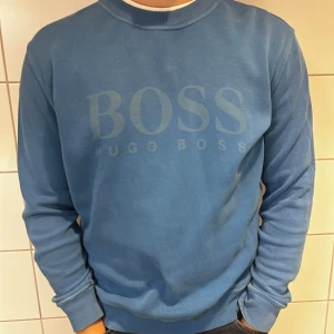 Blå tröja från Hugo Boss - Säljer en snygg blå tröja från Hugo Boss. Den har en stilren design med märkets logga tryckt på framsidan. Tröjan är långärmad och har ribbade muddar vid ärmslut och nederkant. Perfekt för en casual look!