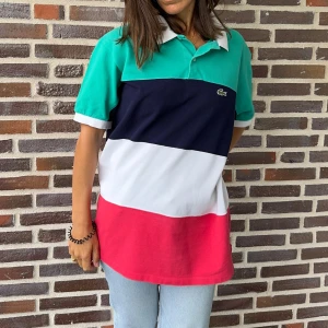 Flerfärgad pikétröja från Lacoste - Säljer en snygg och färgglad Vintage piké polo från Lacoste. Tröjan har breda horisontella ränder i grönt, marinblått, vitt och rött. Den är tillverkad i 100% bomull och har en klassisk krage med knappar. Perfekt för en avslappnad men stilren look! This is perfect for men or women, tenniscore is in fashion 🤩