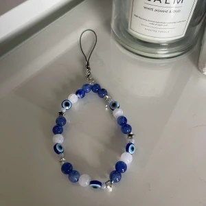 Blått och vitt telefon smycke med ögonmotiv - Säljer en coolt telefon accessoar med blå och vita pärlor och ögonmotiv. Perfekt för att ge en unik touch till din stil. Perfekt accessoar för både vardag och fest!