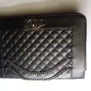 Svart quiltad handväska med kedjedetalj - Säljer en snygg svart handväska i skinn med quiltat mönster. Väskan har en elegant kedjedetalj och en praktisk dragkedja. Perfekt för både vardag och fest!