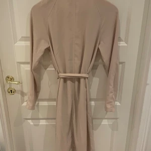 Beige trenchcoat från 157 - Säljer en snygg beige trenchcoat från 157. Den har en klassisk design med ett bälte i midjan för en fin passform. Perfekt för vår och höst! Långärmad och i ett stilrent utförande.