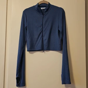 Blå ribbad topp från FB Sister - Säljer en snygg blå ribbad topp från FB Sister. Topp är croppad med dragkedja framtill och har långa ärmar med hål för tummarna. Perfekt för en stilren look!