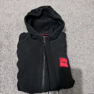 Svart hoodie från Hugo Boss - Säljer en stilren svart hoodie från Hugo Boss med en röd logotyp på bröstet. Hoodien har dragkedja och långa ärmar, perfekt för en casual look. Den är både bekväm och trendig.