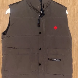 Brun dunväst från Canada Goose - Säljer en brun dunväst från Canada Goose med en röd broderad lönnlöv på bröstet. Västen har en hög krage och stängs med dragkedja och knappar. Två stora fickor framtill ger extra funktionalitet. Perfekt för kyliga dagar!