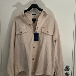 Beige overshirt från GANT - Säljer en snygg beige overshirt från GANT. Skjortan har långa ärmar och två bröstfickor med knappar. Perfekt för lager-på-lager-stil och även inför den kommande våren. Plagget har aldrig använts tidigare och märkeslappen sitter kvar. Nypris är 1800. Hör av er vid intresse:)