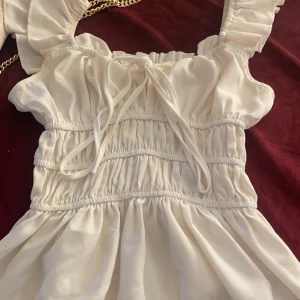 Vit topp med volangdetaljer - Charmig vit topp med volangdetaljer och knytning framtill. Den har en luftig design med breda axelband och elastisk midja för en bekväm passform. Perfekt för sommarens varma dagar.