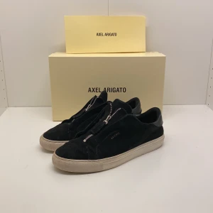 Svarta sneakers från Axel Arigato - Snygga svarta sneakers från Axel Arigato med dragkedja framtill. Skorna har inga stora defekter, förutom smuts, men det är lätt att tvätta bort, skriv vid minsa fundering😃. Ord pris:2500kr