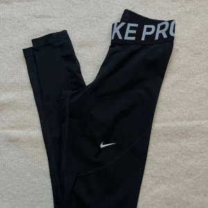 Svarta leggings från Nike - Snygga svarta leggings från Nike med hög midja och meshdetaljer längs benen. Perfekta för träning med en bekväm passform och elastisk midja med logotyp. Tillverkade i ett mjukt och stretchigt material för optimal rörelsefrihet.