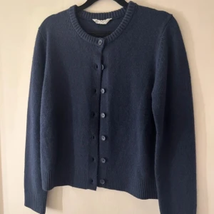 Mörkblå kofta med knappar - Säljer en stilren mörkblå kofta med rund hals och knappar framtill. Koftan har långa ärmar och ribbade muddar vid ärmslut och nederkant. Perfekt för kyliga dagar.
