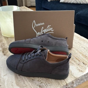 Grå sneakers från Christian Louboutin - Snygga grå sneakers i mocka från Christian Louboutin med den ikoniska röda sulan. Skorna har snörning och en stilren design som passar perfekt för en trendig look. Säljer pågrund av att dem är lite för små. Vet ej om dem är riktiga