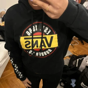 Svart hoodie från Vans - Säljer en svart hoodie från Vans med stort tryck på framsidan i gult och rött. Ärmarna har ett coolt schackrutigt mönster i svart och vitt. Perfekt för en avslappnad stil. 