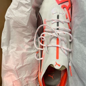 Vita och orange fotbollsskor från Puma - Snygga fotbollsskor från Puma i vitt med orange och svarta detaljer. Skorna har snörning och en modern design som passar perfekt för fotbollsplanen.