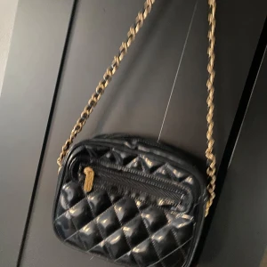 Svart quiltad axelväska med kedja - Snygg svart axelväska med quiltat mönster och guldfärgad kedja. Väskan har en dragkedja framtill och är perfekt för att bära dina nödvändigheter med stil. Den eleganta designen gör den till ett mångsidigt val för olika tillfällen.