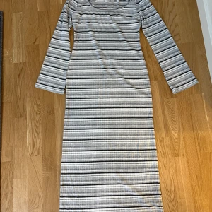 Randig långklänning i ribbat material - Snygg och bekväm långklänning med ränder i svart, grått och vitt från Nelly. Klänningen har en figurnära passform och är tillverkad i ett ribbat material. Den har långa ärmar och en djup rund halsringning, perfekt för både vardag och speciella tillfällen. Endast testad