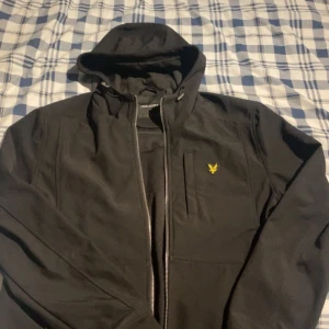 Svart jacka från Lyle & Scott - Säljer en stilren svart jacka från Lyle & Scott med huva och dragkedja. Jackan har en broderad logotyp på bröstet och är perfekt för kyligare dagar. Den är långärmad och har en modern design.