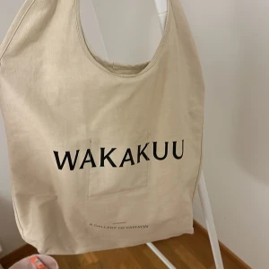 Beige shoppingväska från Wakakuu - Snygg och rymlig beige shoppingväska från Wakakuu med tryckt logotyp på framsidan. Perfekt för att bära med sig allt man behöver under dagen. Väskan har en enkel och stilren design.