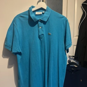 Blå pikétröja från Lacoste - Säljer en klassisk blå pikétröja från Lacoste med den ikoniska krokodilloggan på bröstet. Tröjan har korta ärmar och en traditionell krage med knappar. Perfekt för en stilren sommar outfit använd få tals gånger