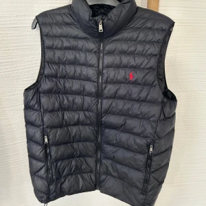 Svart dunväst från Polo Ralph Lauren - Snygg svart dunväst från Polo Ralph Lauren med dragkedja och broderad röd logga på bröstet. Västen har en quiltad design och praktiska fickor med dragkedja. Perfekt för en stilren look. Modellen på bild är 190cm.