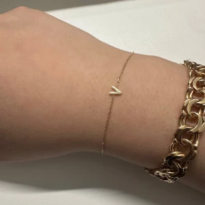 9k Guldarmband med initial V  - Ett elegant armband i 9K guld med en initial V köpt från Ur & Penn. Armbandet är justerbart och kan anpassas till olika handledsstorlekar. Har knappt används så är i ganska nytt och väldigt fint skick. Den köptes den 25e september 2024 och har försäkring 1 års försäkring, alltså fram tills 25e september 2025.   Kvittot skickas med!  Bredd: 5 mm Längd: 18.5 cm Vikt: ca 1 gram