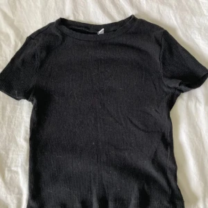 Svart ribbad t-shirt - Säljer en stilren svart ribbad t-shirt med korta ärmar. Perfekt för en enkel och avslappnad look. Passar bra till jeans eller kjol.