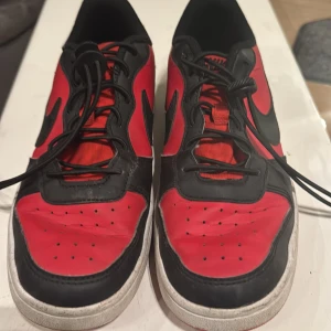 Nike sneakers i rött och svart - Snygga Nike sneakers i rött och svart med klassisk snörning. Skorna har en vit sula och den ikoniska Nike-loggan på sidan och hälen. Perfekta för en sportig look.