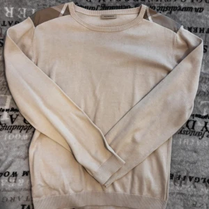 Burberry tröja - Säljer en stilren beige tröja från Burberry med klassiska rutiga detaljer på axlarna. Tröjan har långa ärmar och en rund halsringning. Burberry merinoull i S litet hål kolla sista bilden