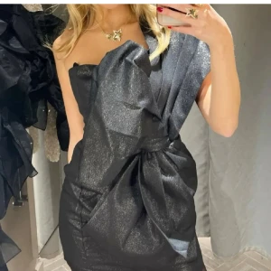 Rosettklänning h&m - Snygg svart klänning med en glittrig finish och en stor rosett framtill. Den är axelbandslös och har en kort längd som ger en elegant look. Perfekt för en kväll ute eller en speciell tillställning🤩Dragkedjan kan vara lite svårt att dra upp vid ett visst ställe men är man försiktig så går det! NY MED PRISLAPP