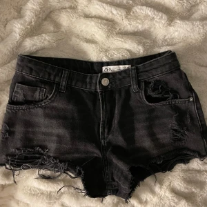 Svarta jeansshorts från Zara - Snygga svarta jeansshorts från Zara med slitna detaljer och fransig kant. Perfekta för en avslappnad stil. De har en klassisk femficksdesign och knappgylf.