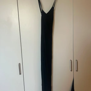Svart långklänning med smala axelband - Elegant svart långklänning med smala axelband och en stilren design. Perfekt för en kväll ute eller en speciell tillställning. Klänningen har en figurnära passform som framhäver silhuetten.
