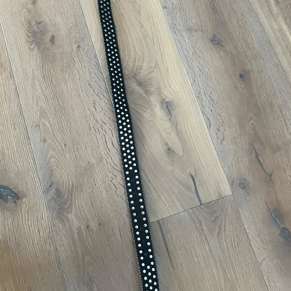 Snyggt svart bälte med glittriga detaljer och nitar. Bältet har en stor spänne dekorerad med svarta stenar och en cool design. Perfekt för att ge din outfit en extra edge.. Muu.