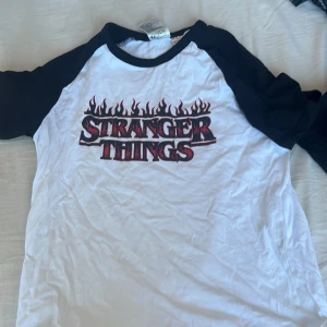 Vit och svart Stranger Things t-shirt från H&M - Cool vit t-shirt med svarta ärmar från H&M med Stranger Things-tryck i rött och svart. Perfekt för fans av serien! Tillverkad i bomull för en skön känsla.
