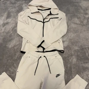 Vit träningsdräkt från Nike - Snygg vit träningsdräkt från Nike med svarta detaljer. Setet inkluderar en huvtröja med dragkedja och matchande byxor med dragsko i midjan. Perfekt för en sportig look.