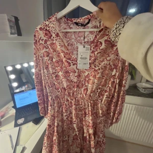 Blommig klänning från Zara - Säljer en vacker blommig klänning från Zara i rosa och vit. Klänningen har långa ärmar och en v-ringad design. Perfekt för vår och sommar med sitt lätta och luftiga material.