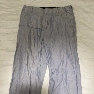 Grå kostymbyxor från H&M - Snygga grå kostymbyxor från H&M med diskret mönster. Byxorna har en klassisk passform med knappar och hällor för bälte. Även inbyggt resår i midjan så man kan justera storleken. Byxorna kan självklart strykas innan köp.