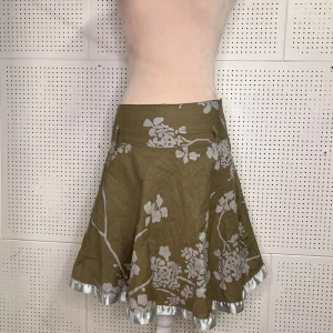 Beige blommig kjol med satin detaljer - Charmig midi kjol med vitt blommönster och en glansig satin kant. Kjolen har en utsvängd form som ger en fin siluett. Perfekt för en somrig look!