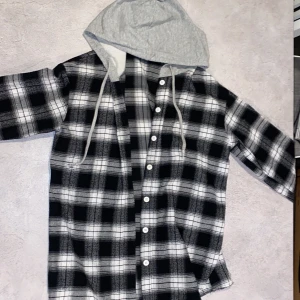 Svartvit rutig skjorta med grå huva - Säljer en cool svartvit rutig skjorta med grå huva och knappar framtill. Skjortan har långa ärmar och en avslappnad stil. Perfekt för en casual men vågad look.