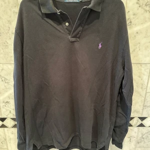 Svart pikétröja från Ralph Lauren - Säljer en klassisk svart pikétröja från Ralph Lauren med lila broderad logga på bröstet. Tröjan är långärmad och tillverkad i 100% bomull, vilket gör den både bekväm och stilren. Perfekt för en avslappnad men ändå elegant look.