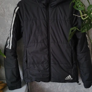 Svart dunjacka från Adidas - Säljer en svart dunjacka från Adidas med klassiska vita ränder på ärmarna. Jackan har en dragkedja framtill och en huva för extra skydd mot väder. Perfekt för kyligare dagar. Adidas-loggan syns tydligt på framsidan. 🖤