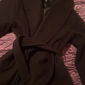Svart kappa från Vero Moda - Snygg svart kappa från Vero Moda i storlek M. Den har en elegant design med knytbälte i midjan och långa ärmar. Perfekt för kyligare dagar. Till våren/hösten, köpt för 800kr och inte använd mer än ca 4 gånger då den är för stor 