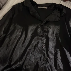 Svart blus med knappar - Snygg svart blus med knappar framtill. Den har en glansig finish och en klassisk krage. Perfekt för en stilren look.