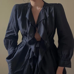Vintage DKNY Linen Black Shirt - DKNY Black Open Ruffle Woven Linen Top with a Silk Grosgrain Ribbon Tie at the Waist. Cinched Waist with Decorative Trims. 3/4 sleeve with Buttoned Sleeves.  Can be worn loosely fitted as a layering top or a black bralette for a more intimate look. Original Tag Size 4. Model is 160cm (5”3) and generally fits S/M. Excellent Condition.  67 CM/ 26.4 IN Length 50 CM/ 19.7 IN Sleeve 38 CM/ 15 IN Shoulders 84 CM/ 33.1 IN Chest 76 CM/ 29.9 IN Waist 