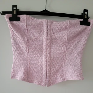 Ljusrosa korsett - Säljer en söt ljusrosa korsett med ett unikt texturerat mönster. Den har en praktisk hyskknäppning framtill och detaljer längst upp. Oanvänd storlek S från HM