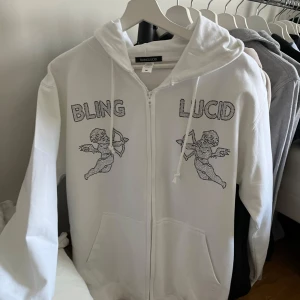 Vit hoodie från Bling Lucid - Alldrig använd Säljer en vit hoodie från Bling Lucid med dragkedja och huva. Den har ett coolt tryck med texten 'BLING LUCID' och två änglar i strass på framsidan. Perfekt för en avslappnad stil med lite extra bling.