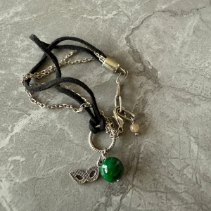 Armband med grön pärla och silverdetaljer - Unikt armband med en grön pärla och silverfärgade detaljer. Armbandet har en kombination av svart band i äkta mocka och silverkedja, samt en dekorativ lövberlock. Perfekt för att ge en personlig touch till din stil.